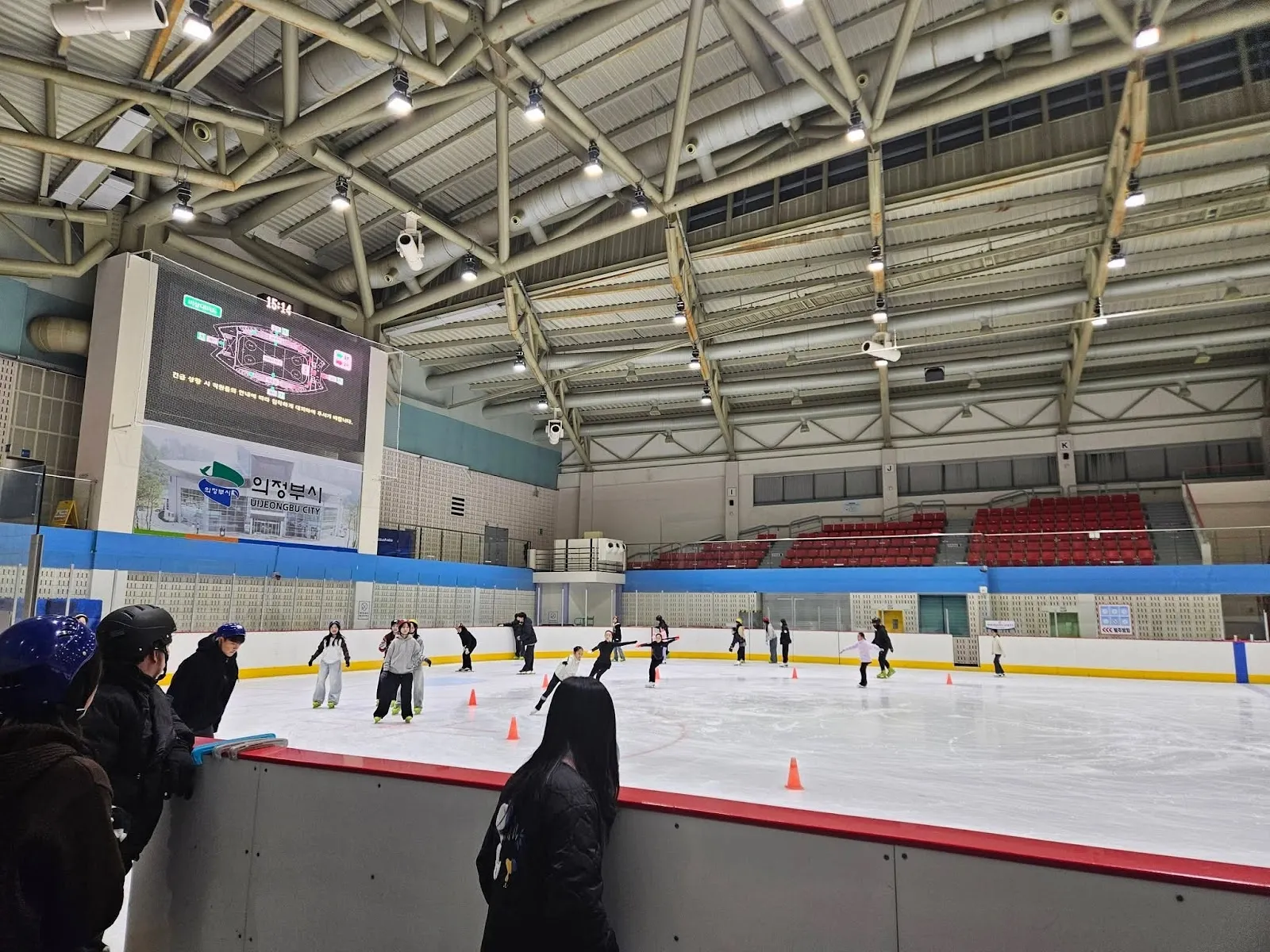 A game on the ice of Uijeongbu Indoor Ice Rink, seen from the stands across the sheet with the video screen and the steel roof behind. — Uijeongbu Indoor Ice Rink