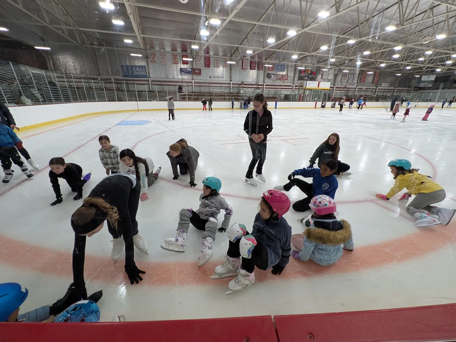 A learn-to-skate group seated on the ice around a faceoff circle with an instructor bending among them, rows of lights under the steel roof and the far boards behind. — Ulin Memorial Rink