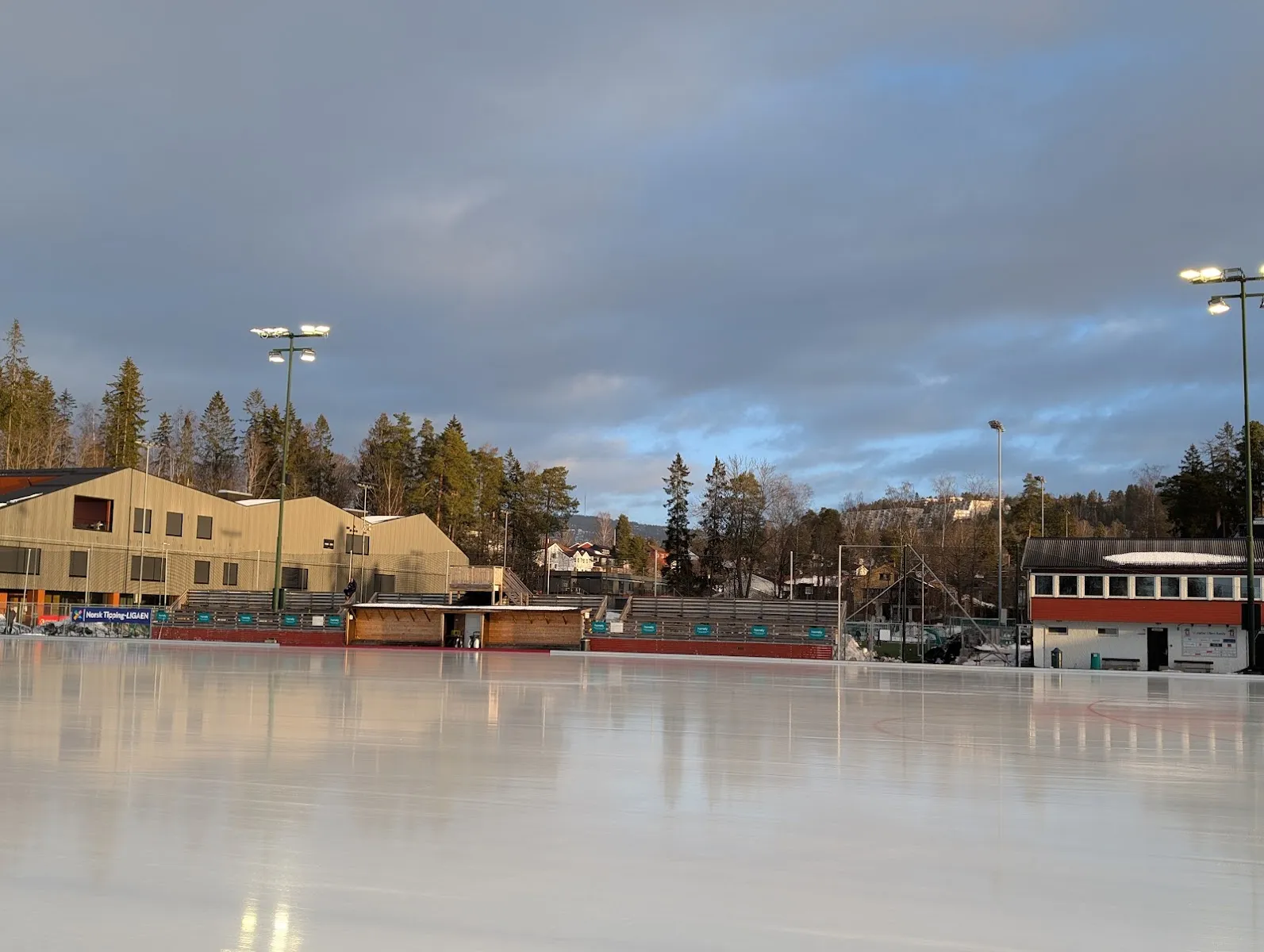 Ullerns kunstis i skumringen, tom, med klubbhuset og flomlysene bak banen. — Ullern kunstisbane