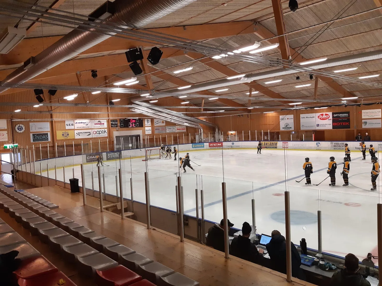 En grupp skridskoåkare på isen i Ulricehamns ishall, med trälåktare och sponsorsargar längs långsidan under trätaket. – Ishallen Ulricehamn