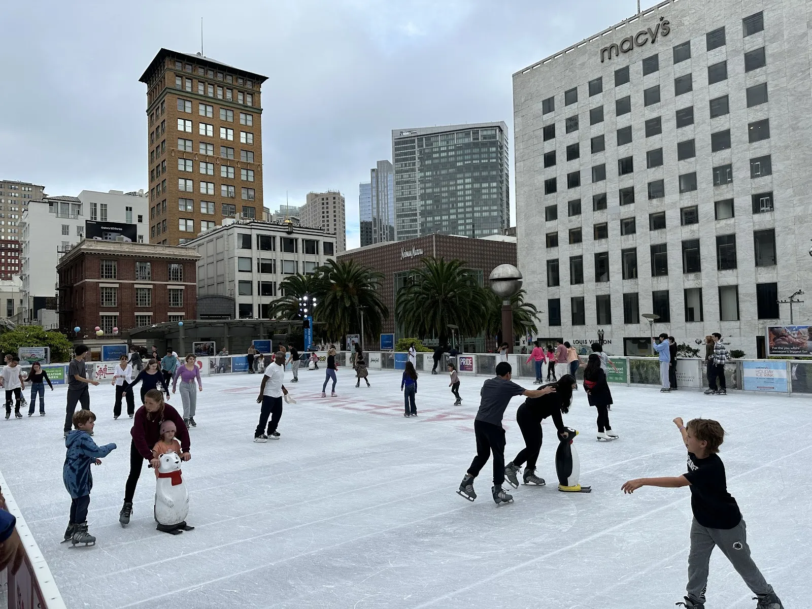 The outdoor ice looking across the plaza toward the department store and office towers, with skaters spread across the sheet. — Union Square Ice Rink