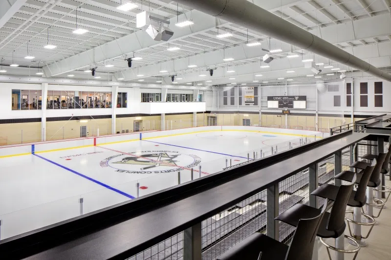 The sheet with a Penguins crest at centre seen from a raised counter along the glass, a bright steel-trussed hall with a long window wall behind. — UPMC Lemieux Sports Complex