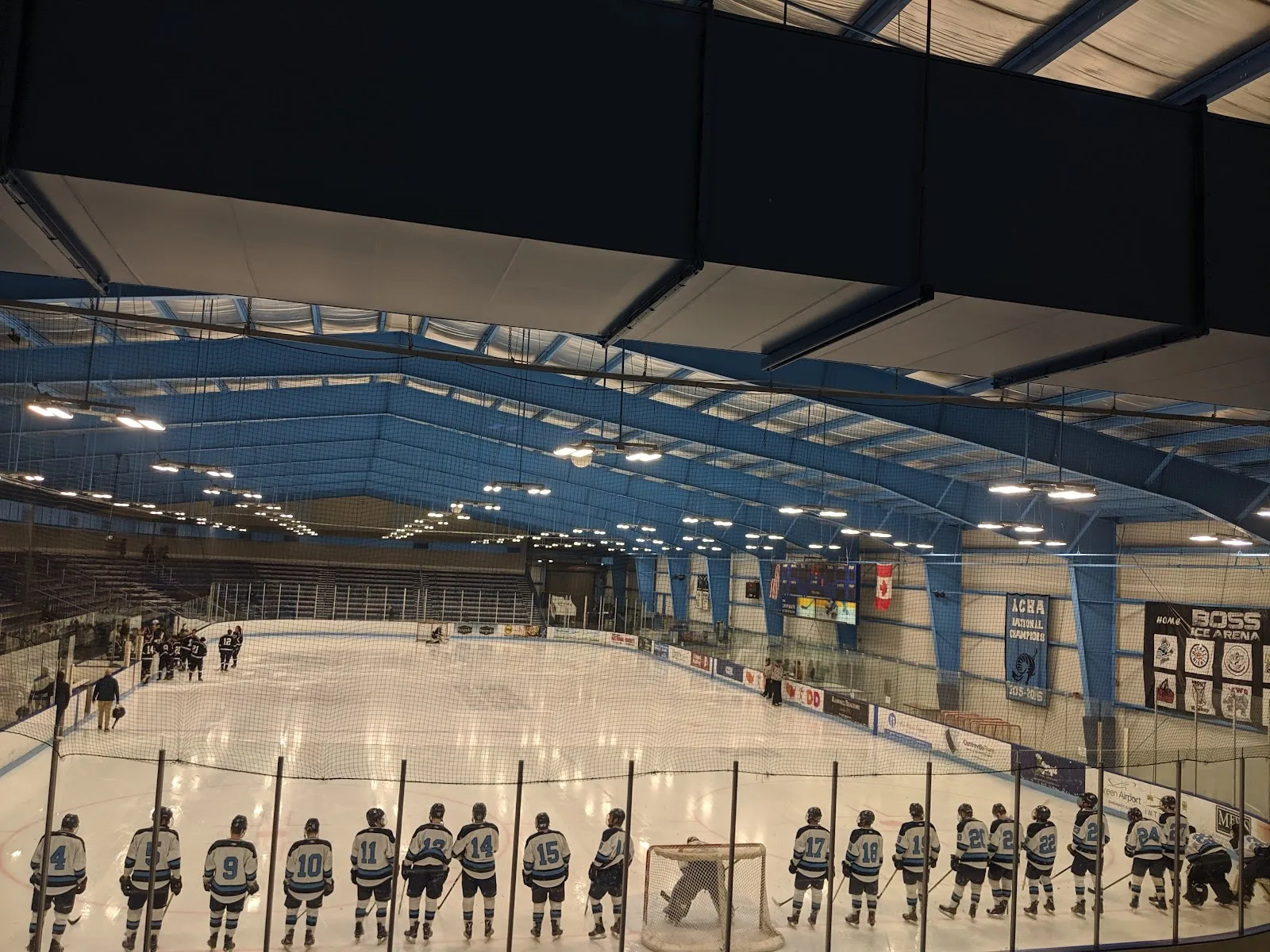 Un partido visto desde las butacas bajas, con jugadores repartidos por el hielo y un equipo alineado a lo largo de las vallas del fondo bajo un techo de acero azul. — URI Boss Arena