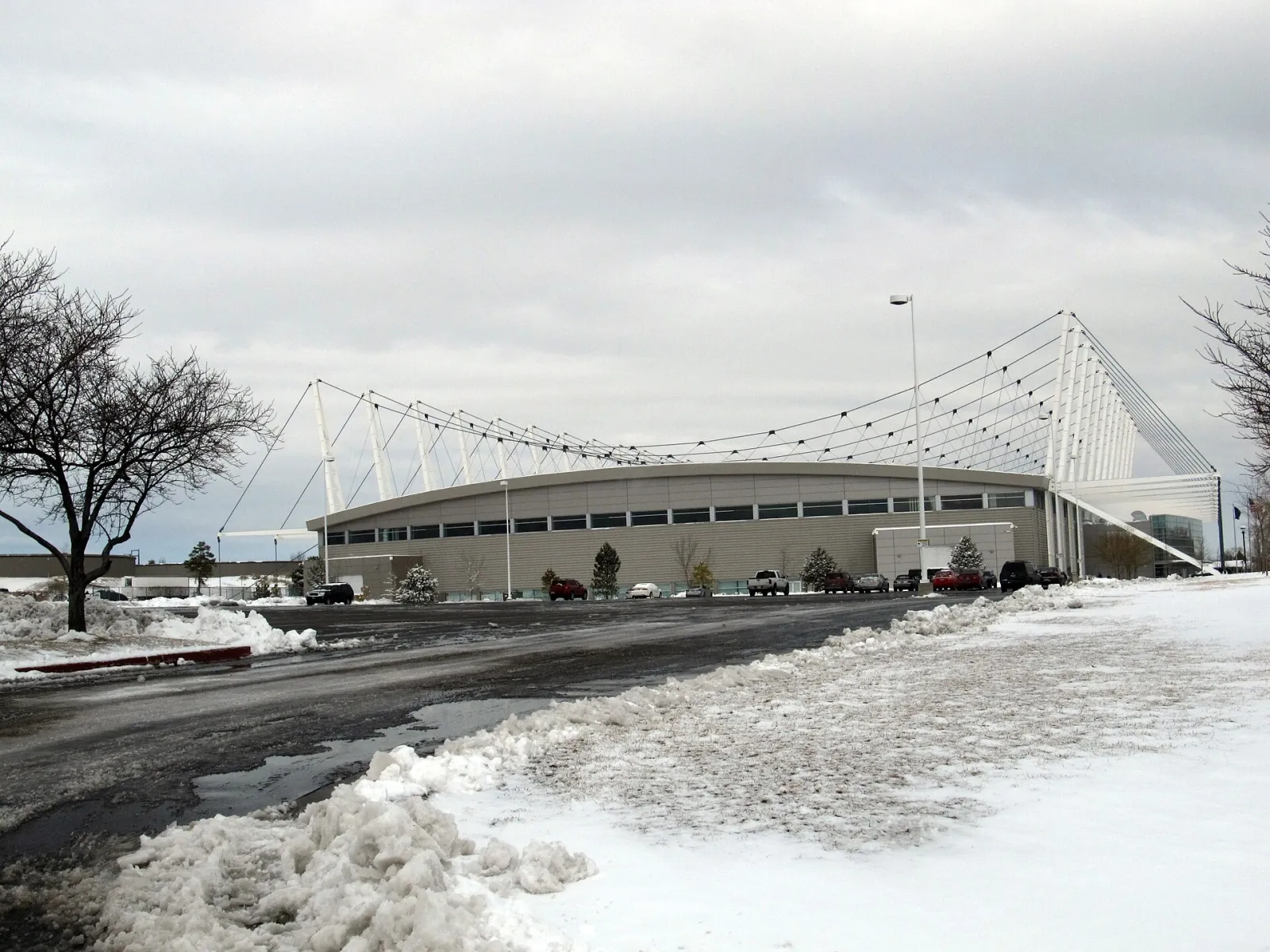 The long, low Oval building with its row of white masts and the suspension cables that carry the roof, seen across a snowy parking lot. — Utah Olympic Oval
