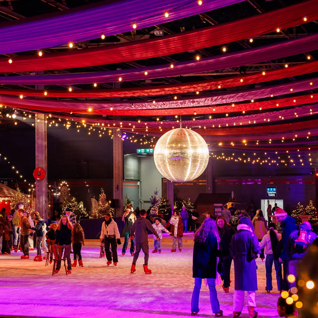 Schaatsers op de overdekte ijsbaan van het Utrechtse Winterspektakel onder rode plafonddraperieën en een discobal; schaatsers op afstand. – Het Utrechtse Winterspektakel