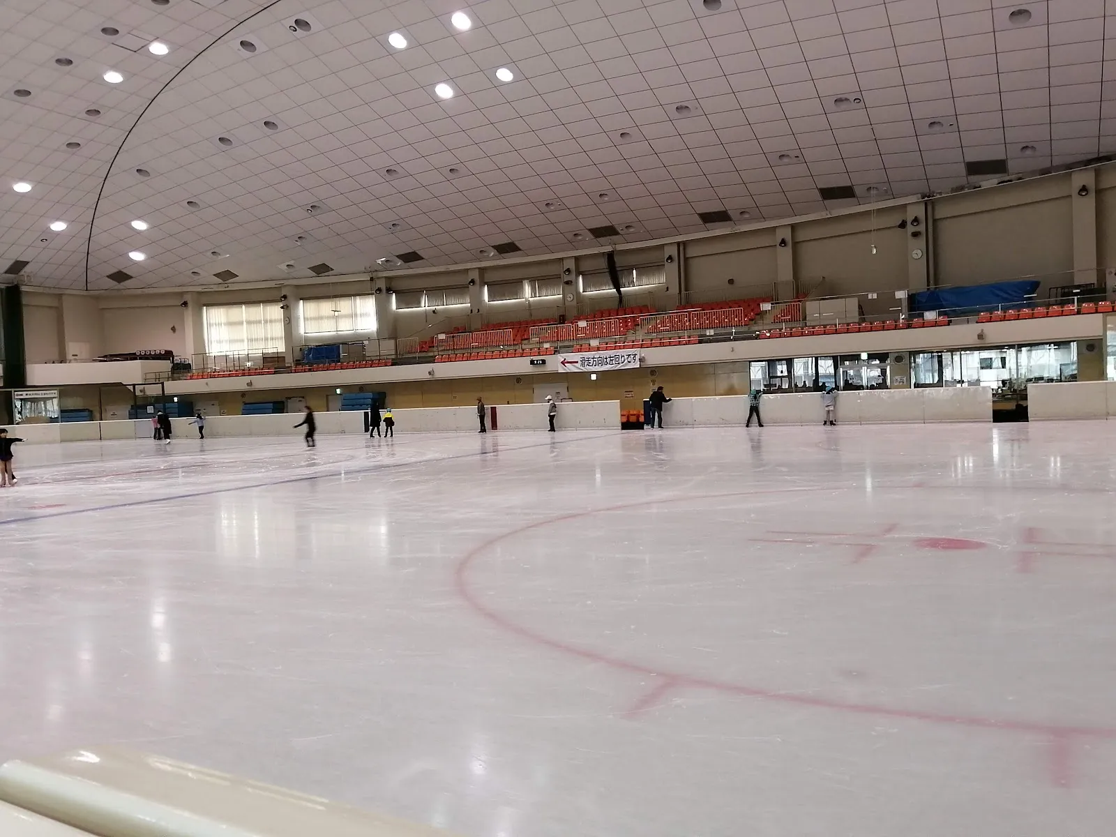 The ice sheet of the Utsunomiya City Skate Center from the rink side, with a few small skaters far across the ice and the stands beyond — Utsunomiya City Skate Center