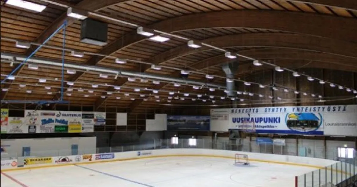 The empty ice sheet of Uudenkaupungin jäähalli seen from the stands, under a curved timber roof with rows of lights and advertising boards along the far side. – Uudenkaupungin jäähalli