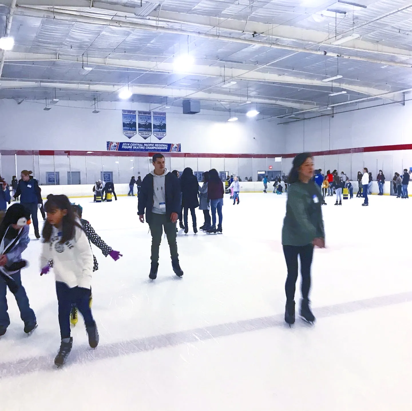 A public skating session on the white sheet, skaters spread across the ice with Central Pacific regional championship banners and a pale steel roof behind them. — Vacaville Ice Rink