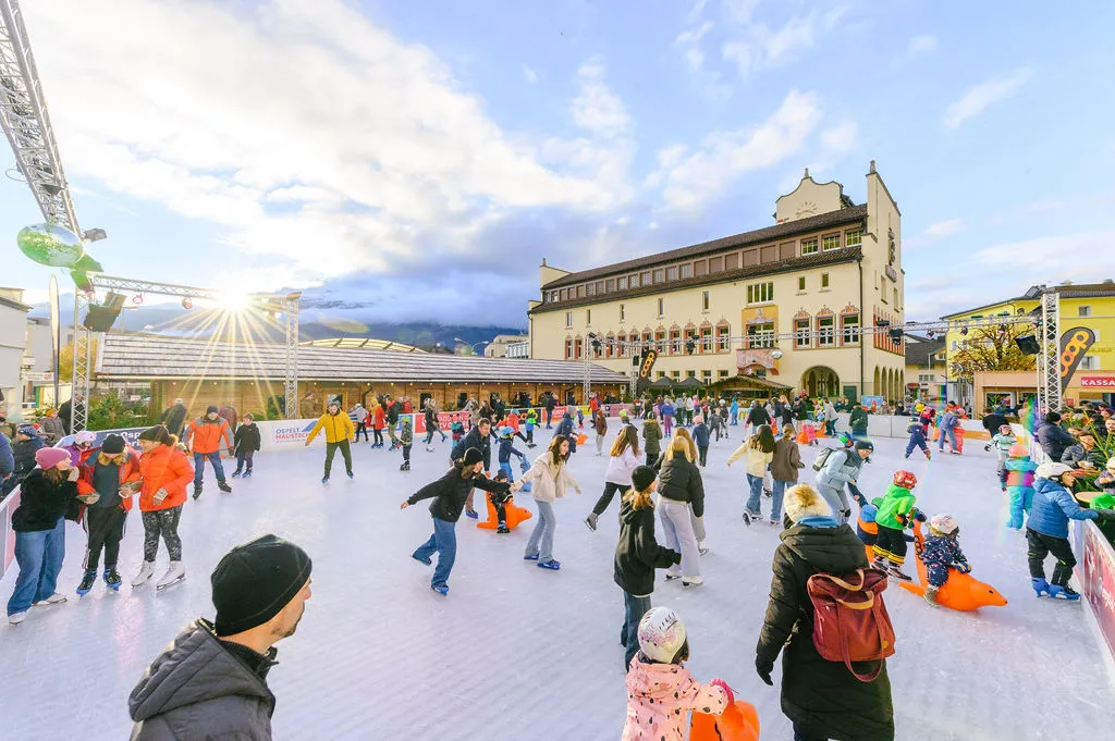 Das Eisfeld auf dem Rathausplatz in Vaduz vor dem Rathaus an einem sonnigen Tag, mit den Markthäuschen entlang der linken Seite – Vaduz on Ice