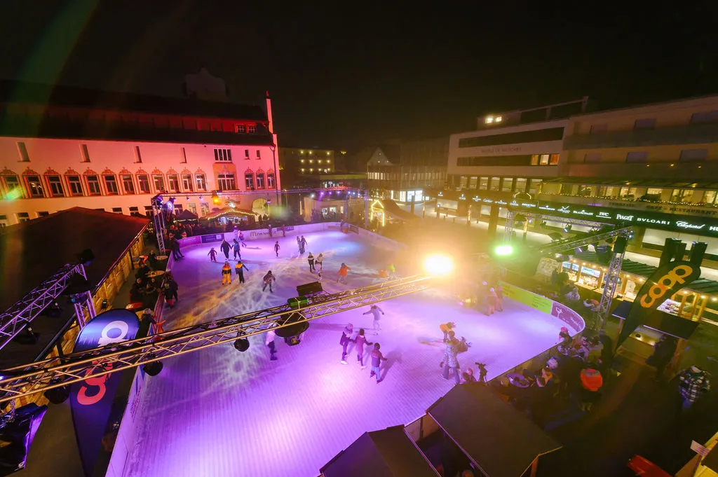 Das ganze Eisfeld bei Nacht von oben gesehen, violett beleuchtet, mit Schlittschuhläufern und dem Rathaus im Hintergrund – Vaduz on Ice