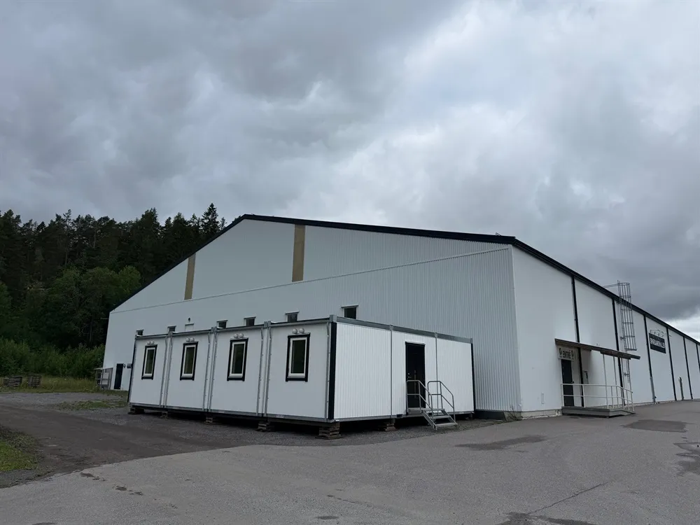 The white steel-clad hall building from the car park, with the rows of lodging containers built against its front wall. – Valdemarsviks Ishall