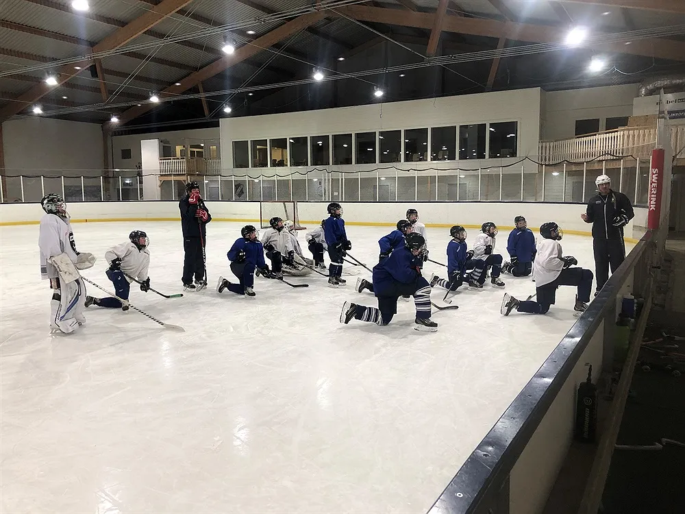 The ice sheet of Valdemarsviks ishall during a youth hockey practice, seen from the players' box under a timber-trussed roof with the cafeteria windows above the boards. – Valdemarsviks Ishall