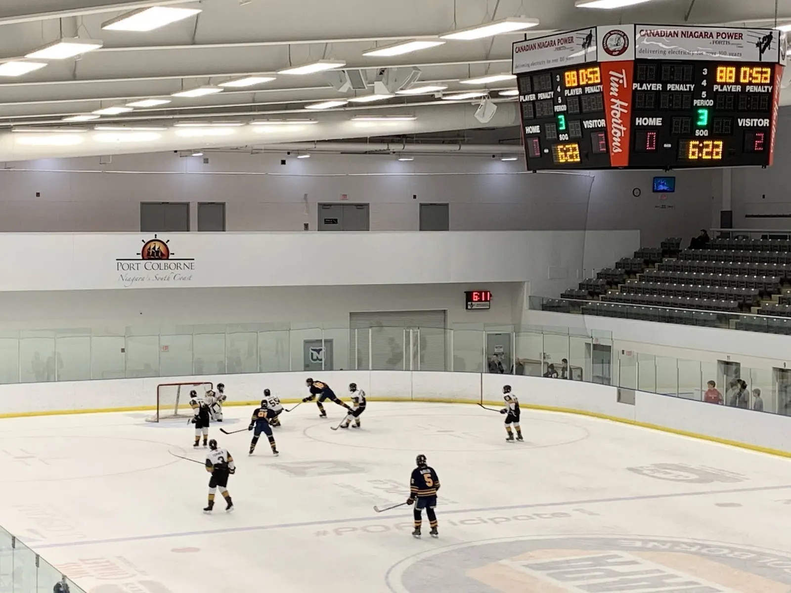 A game seen across the sheet under a long arched roof, players spread near the far net below a score clock, a grandstand along the right. — Vale Health & Wellness Centre