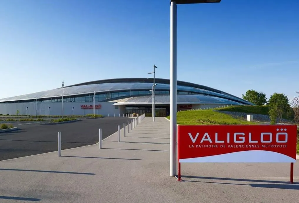 Le bâtiment bas à toit courbe vu de l’autre côté du parvis pavé en pleine lumière, avec une enseigne rouge portant VALIGLOÖ La patinoire de Valenciennes Métropole au premier plan. — Valigloo