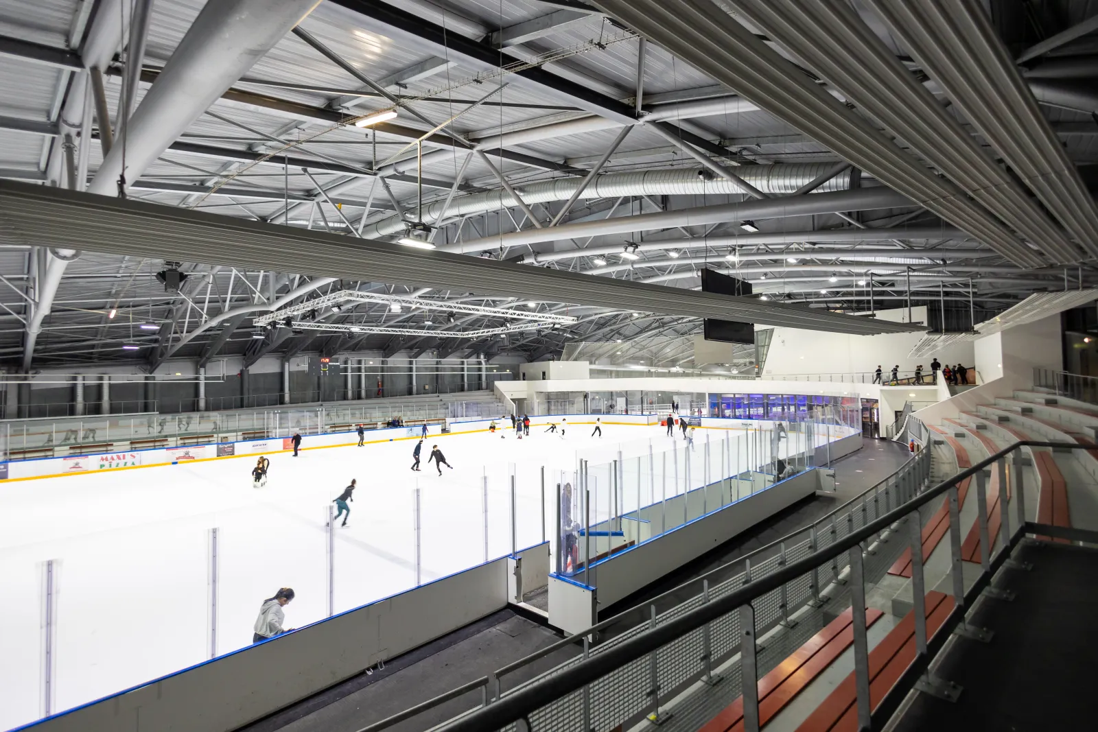 La piste olympique vue des gradins : glace blanche avec quelques patineurs au fond, bandes et vitres à sommet bleu, sièges rouges le long des bandes et charpente à fermes d’acier. — Valigloo