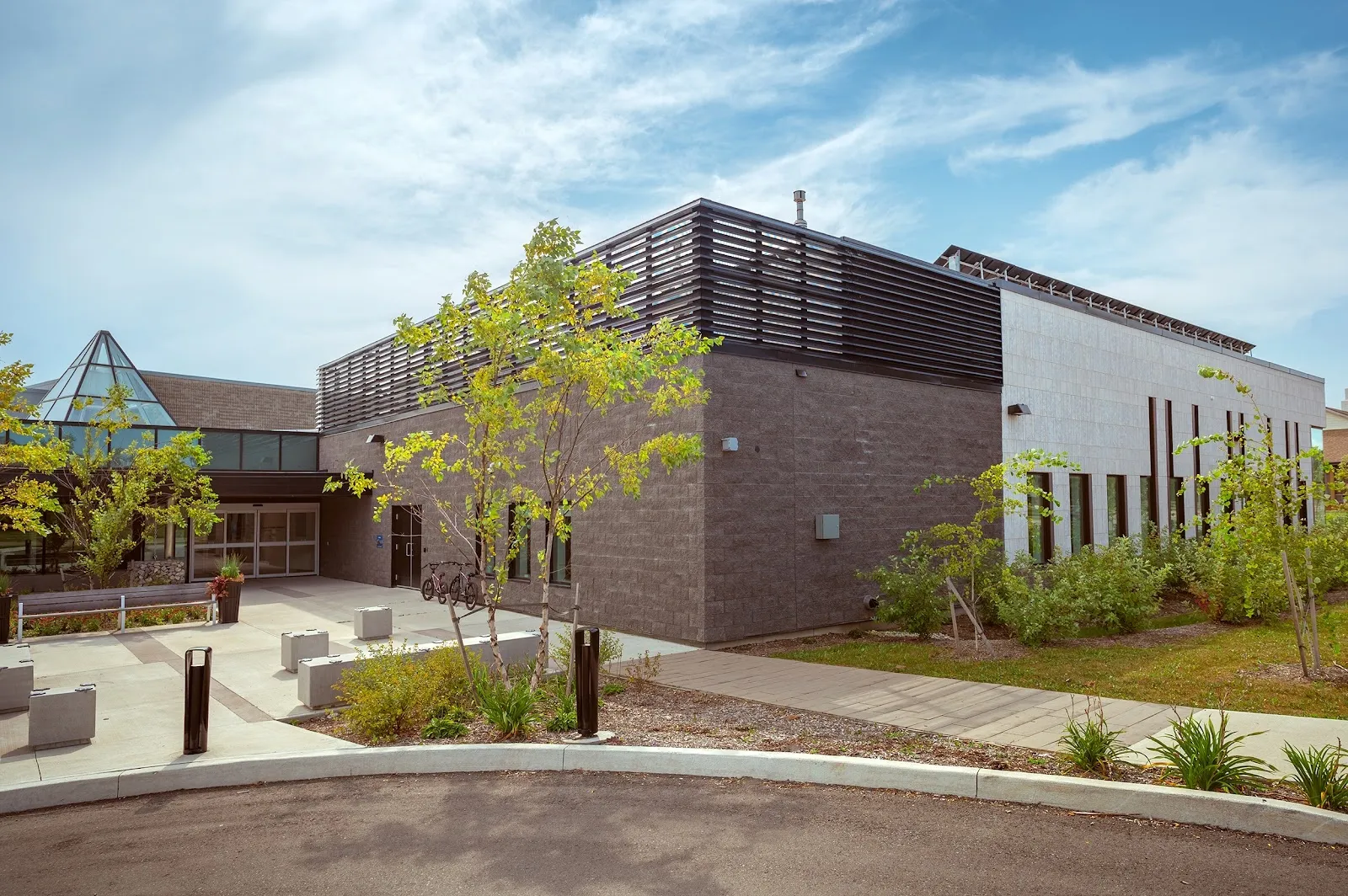 The Valley Park Community Centre's modern brick and timber-clad wing with a glazed corner, young trees and a paved courtyard in front under a pale blue sky. — Valley Park Arena