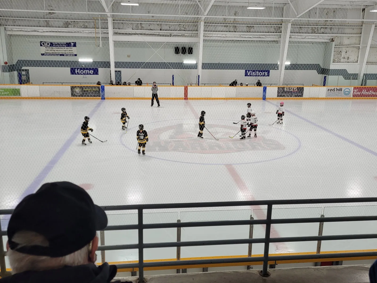 A game seen from the stands with the far end of the sheet and a faceoff circle, players small across the ice under a white steel-trussed roof. — Valley Park Arena