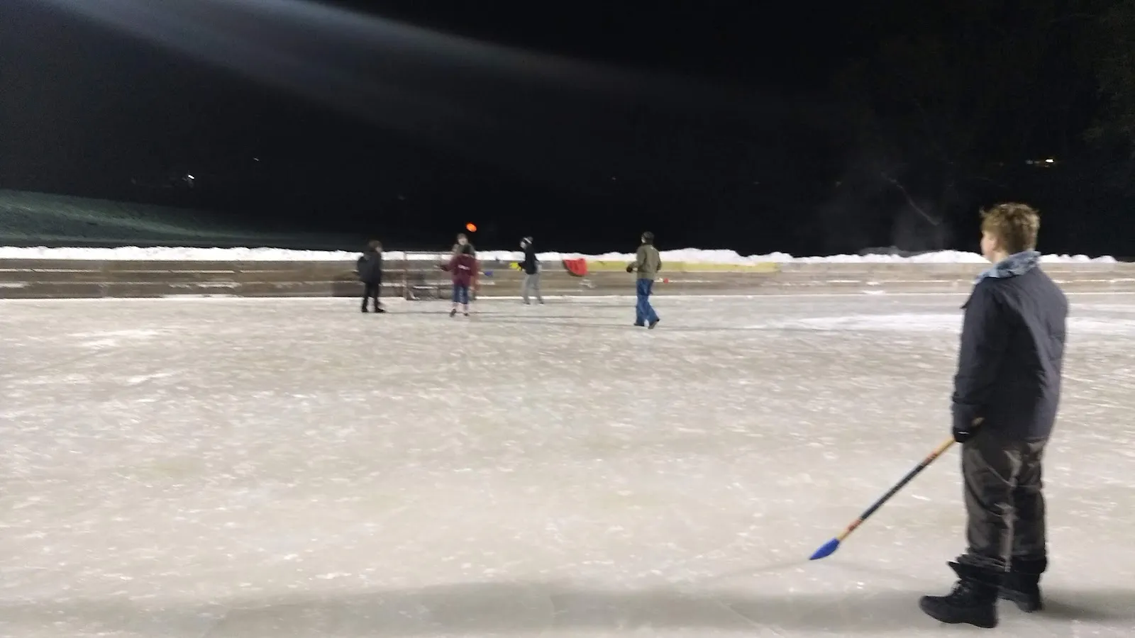 A pickup hockey game on the lit outdoor ice at Valley Place Park at night, with players across the sheet in the distance and a person with a stick standing at the edge of the ice. — Valley Place Park Ice Rinks