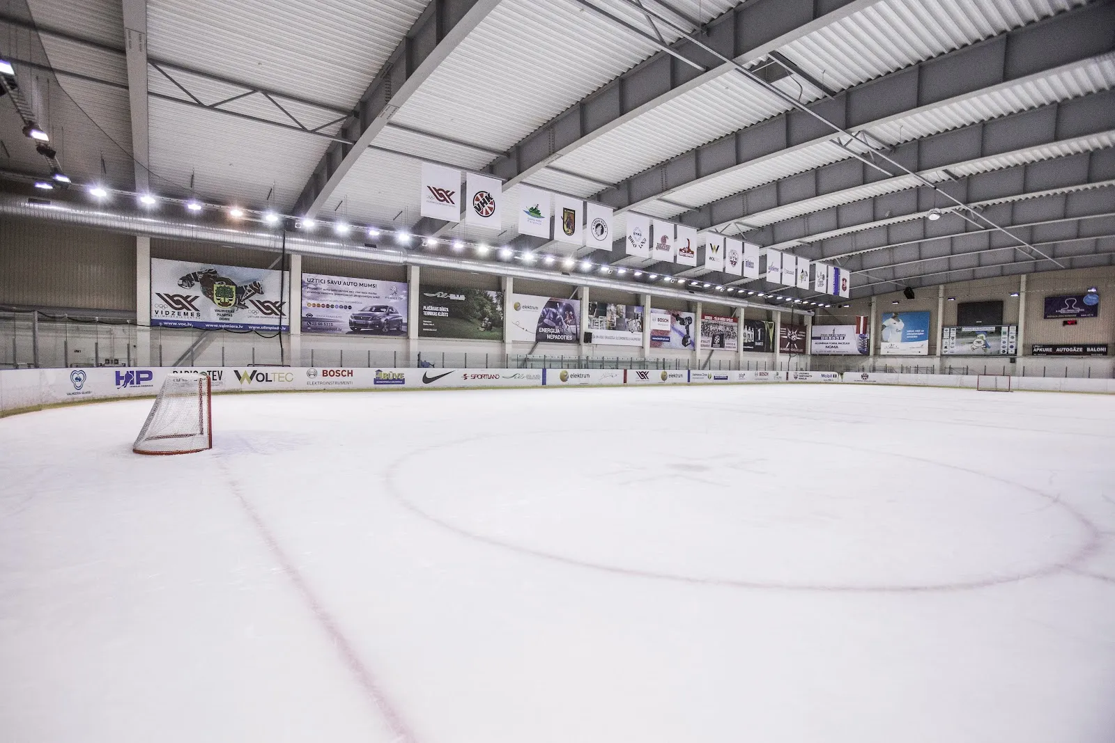 The empty ice sheet of the Valmiera Olympic Centre's KIA ice hall, with the sponsor boards along the boards and the steel roof above — Valmieras Olimpiskā centra ledus halle
