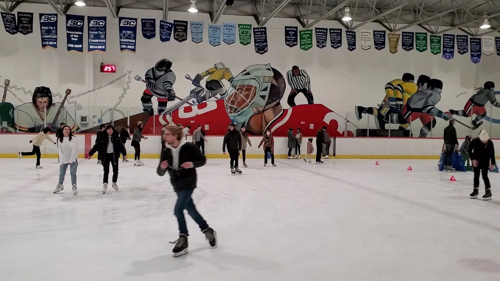 A public session under a steel-trussed roof, skaters spread across the sheet below a mural wall of players and a row of championship banners. — Britannia Rink