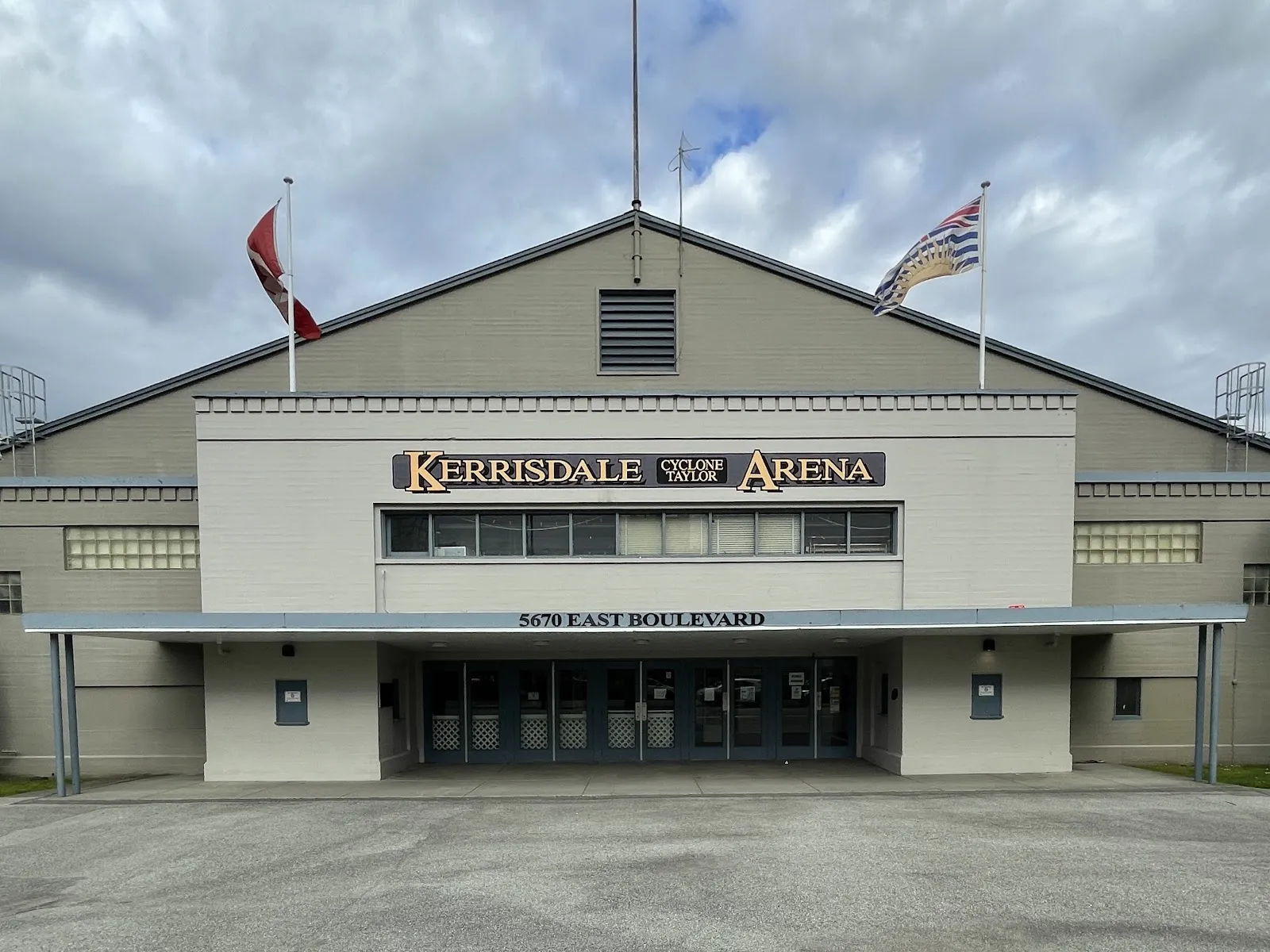 Front facade of Kerrisdale Cyclone Taylor Arena with a large gold-lettered 'KERRISDALE CYCLONE TAYLOR ARENA' sign and '5670 EAST BOULEVARD' address over the entrance canopy, Canadian and BC flags flying above. — Kerrisdale Cyclone Taylor Arena