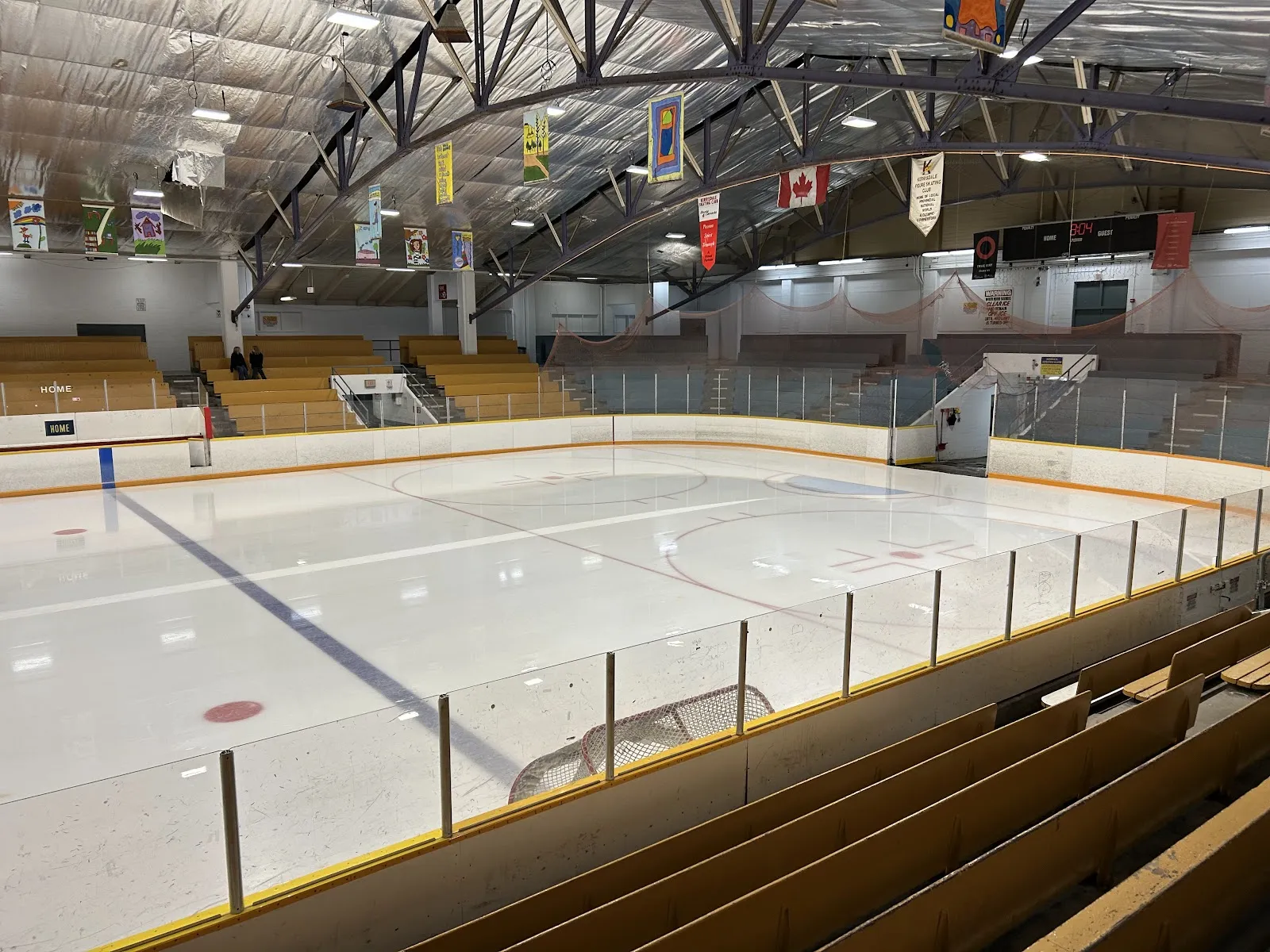 Empty ice sheet at Kerrisdale Cyclone Taylor Arena seen from the stands, with wooden bleachers, a 'Kerrisdale Figure Skating Club' banner and a Canadian flag hanging from the arched roof trusses. — Kerrisdale Cyclone Taylor Arena
