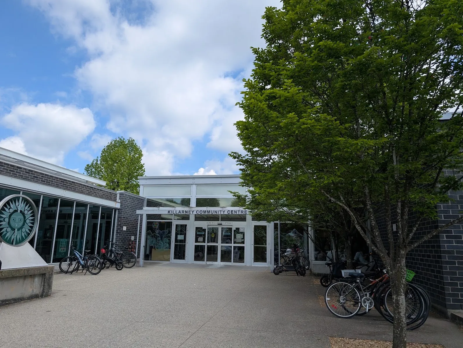The Killarney Community Centre entrance at 6260 Killarney Street, its name over the glass doors, with bikes racked under a tree. — Killarney Rink