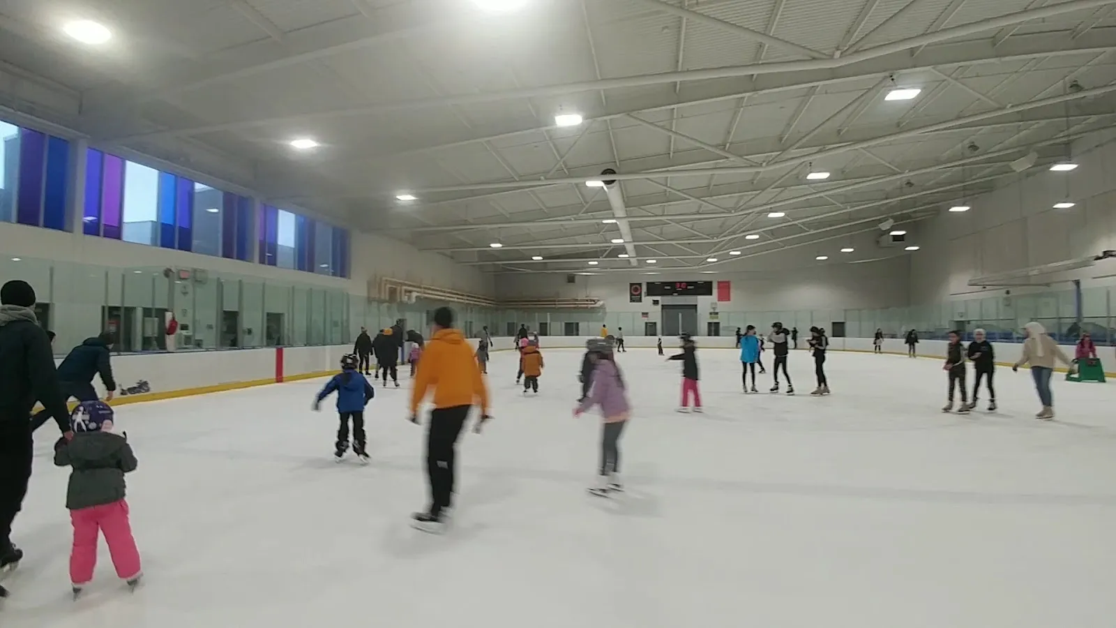 A public session on a wide sheet, skaters spread across the ice under a steel-braced roof with a blue-lit scoreboard strip on the far wall. — Killarney Rink
