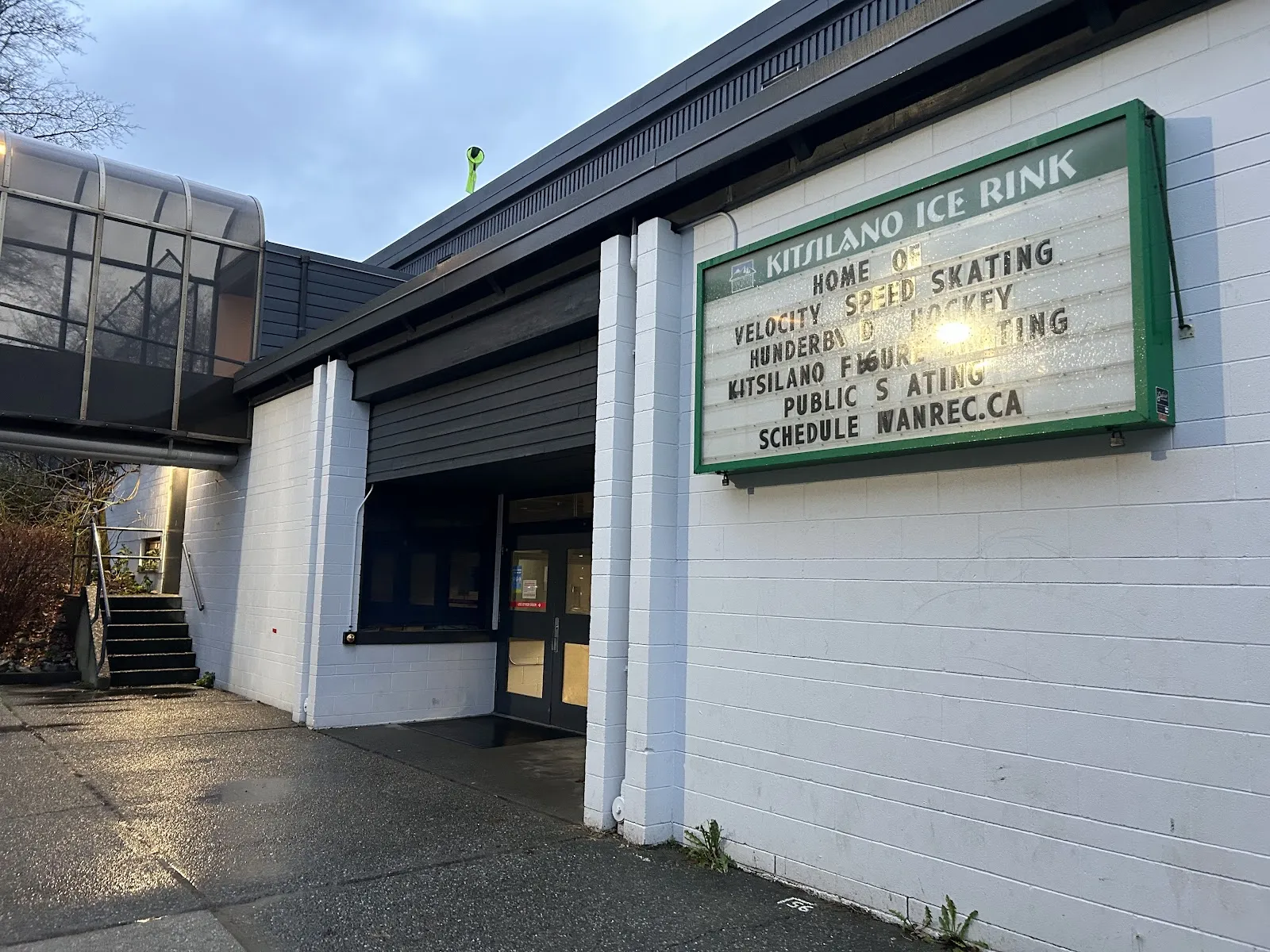 Building entrance with an illuminated sign reading "Kitsilano Ice Rink – Home of Velocity Speed Skating, Thunderbird Hockey, Kitsilano Figure Skating, Public Skating" beside the covered walkway bridge. — Kitsilano Rink