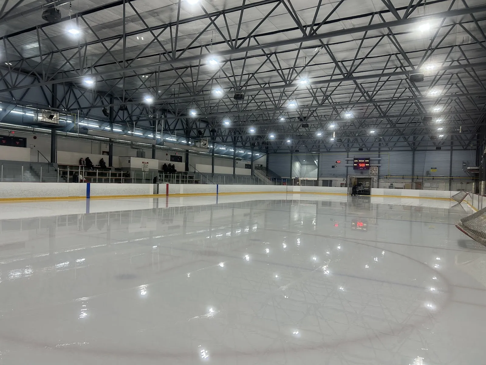 Empty ice sheet and steel-trussed arena interior with bleachers, scoreboard and net visible; no skaters on the ice. — Kitsilano Rink