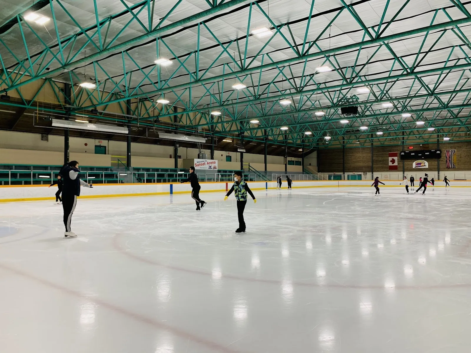 Skaters spread across a wide sheet under a green steel-arched roof, red and blue lines on white ice and a long wall behind the boards. — Sunset Rink