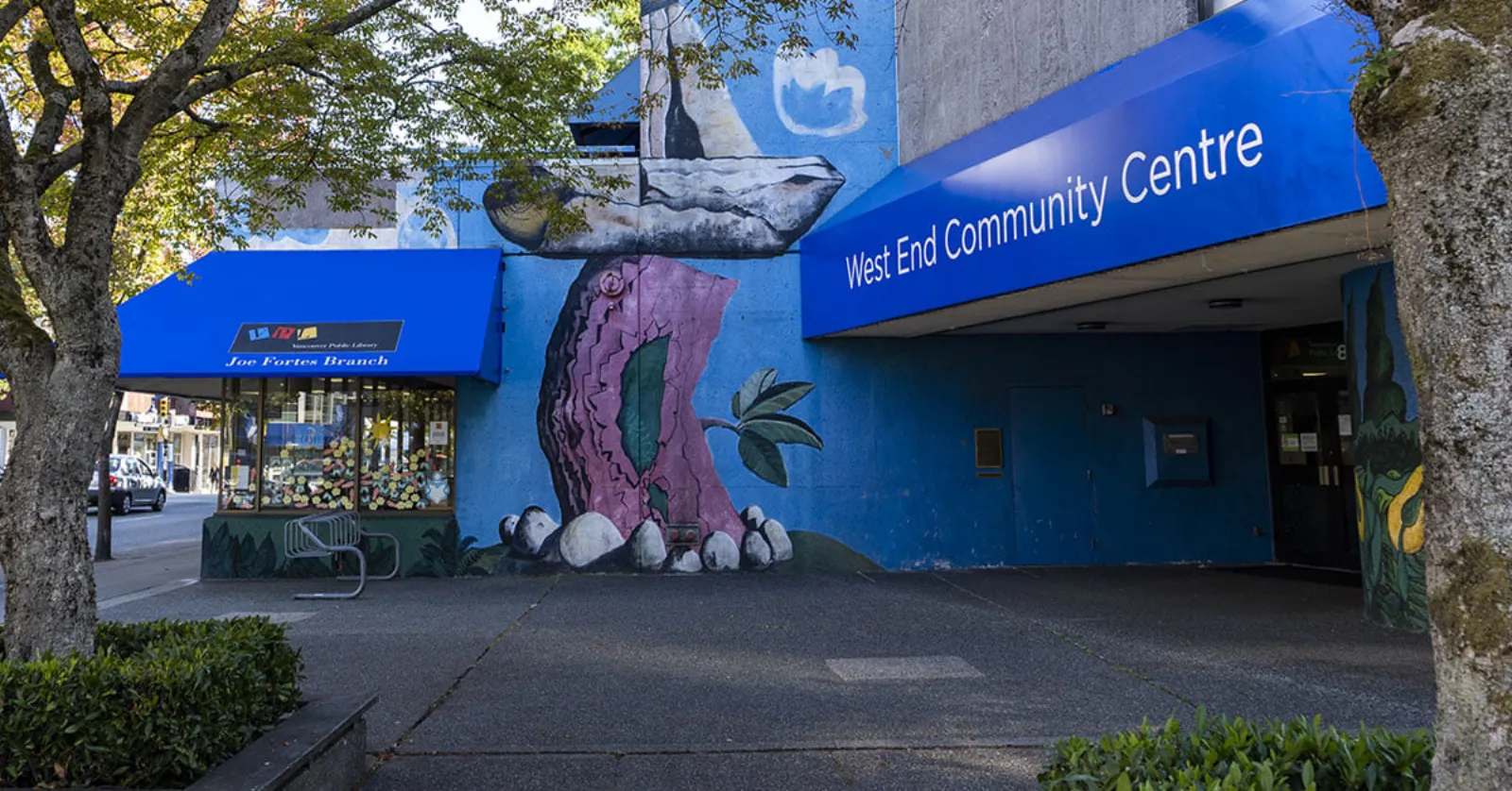 Vista a nivel de la calle de la entrada del West End Community Centre, con un gran toldo azul con el letrero "West End Community Centre", un mural en el muro de concreto y el local contiguo de la sucursal de la biblioteca Joe Fortes. — West End Rink