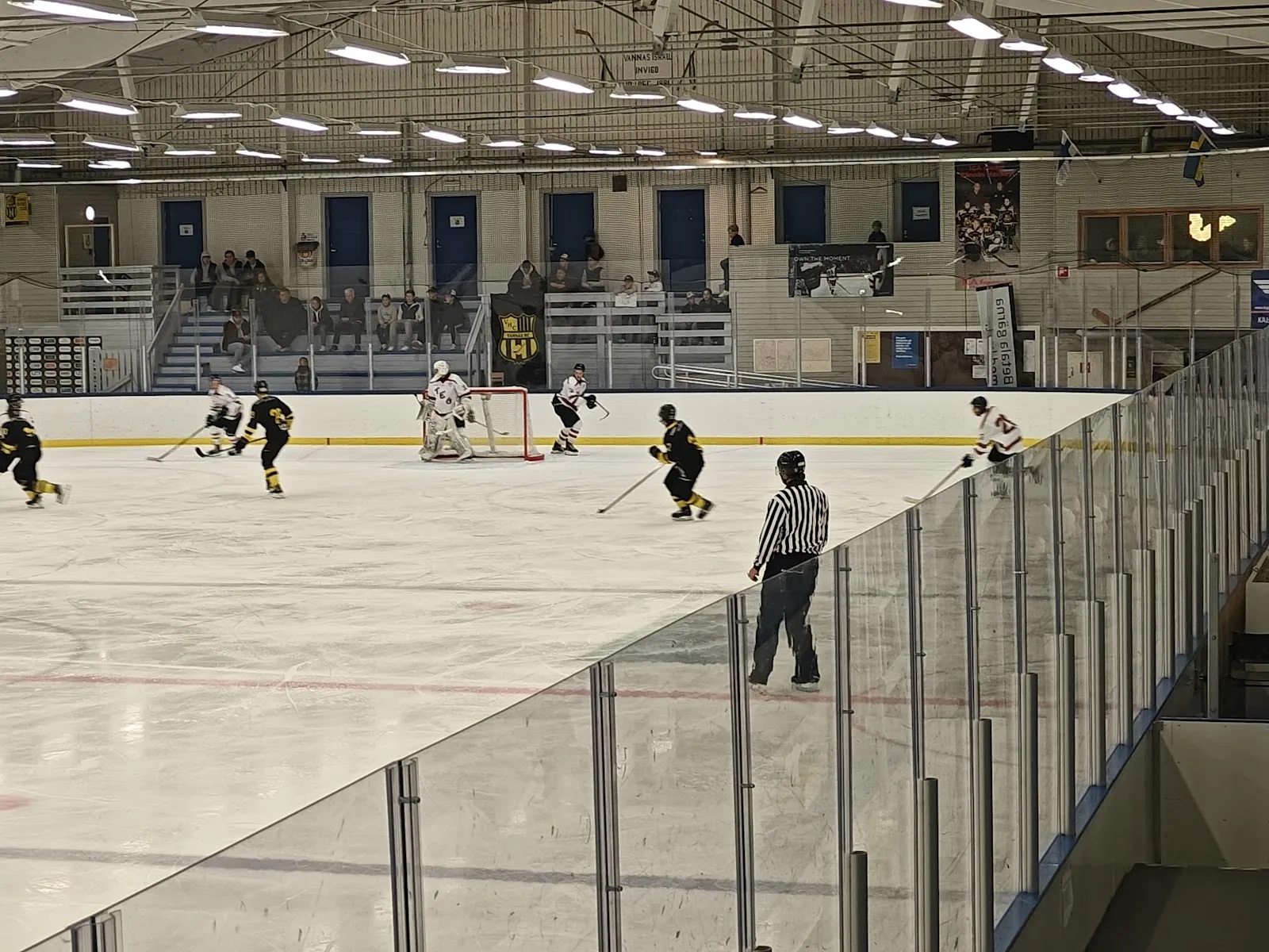 A game on the ice at Vännäs ishall, with the plexiglass along the boards, the roof lighting and the long grandstand behind. — Vännäs ishall