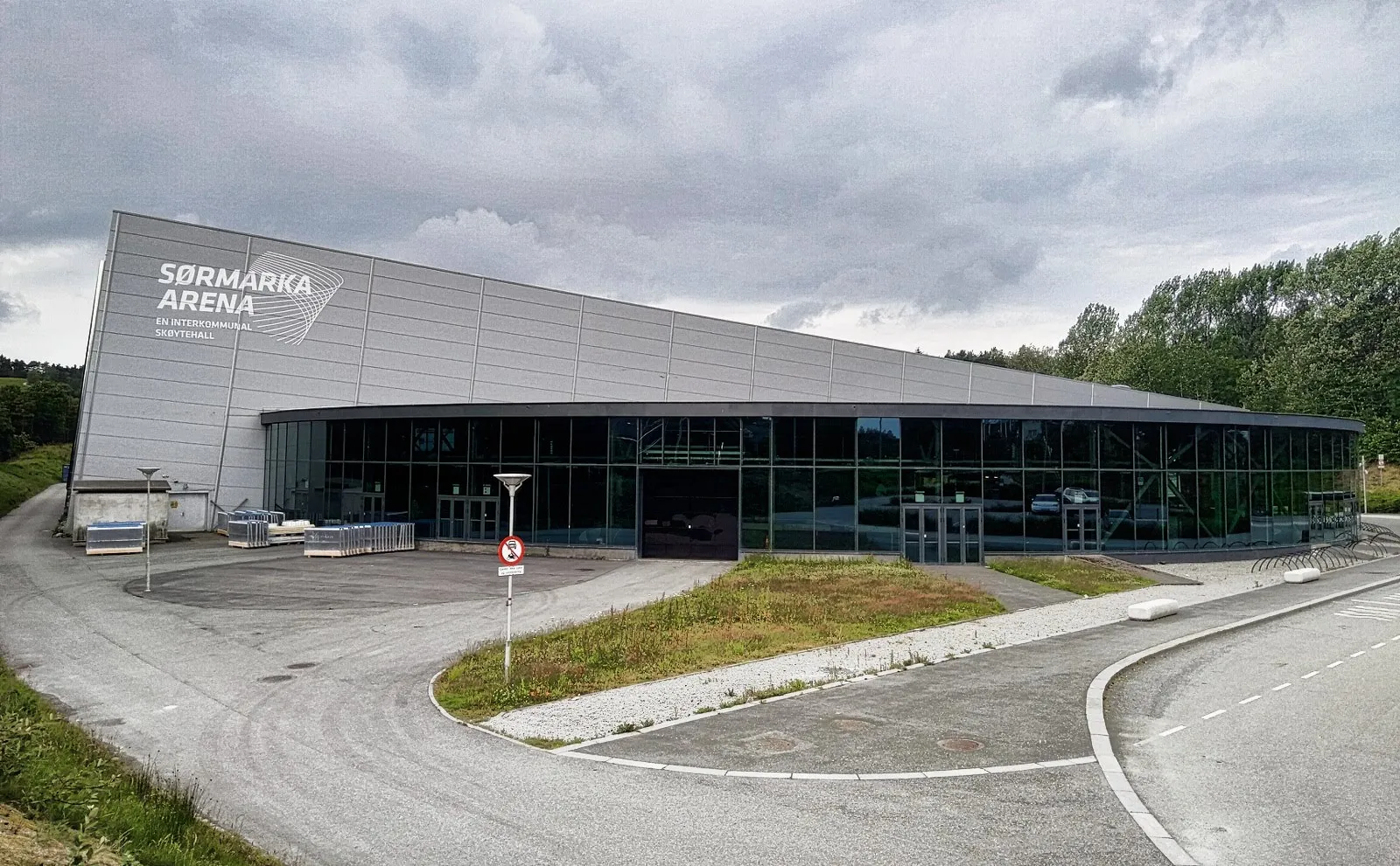 The grey Sørmarka Arena building with its glazed front, seen across the empty car park and access road. — Vår Energi Arena Sørmarka