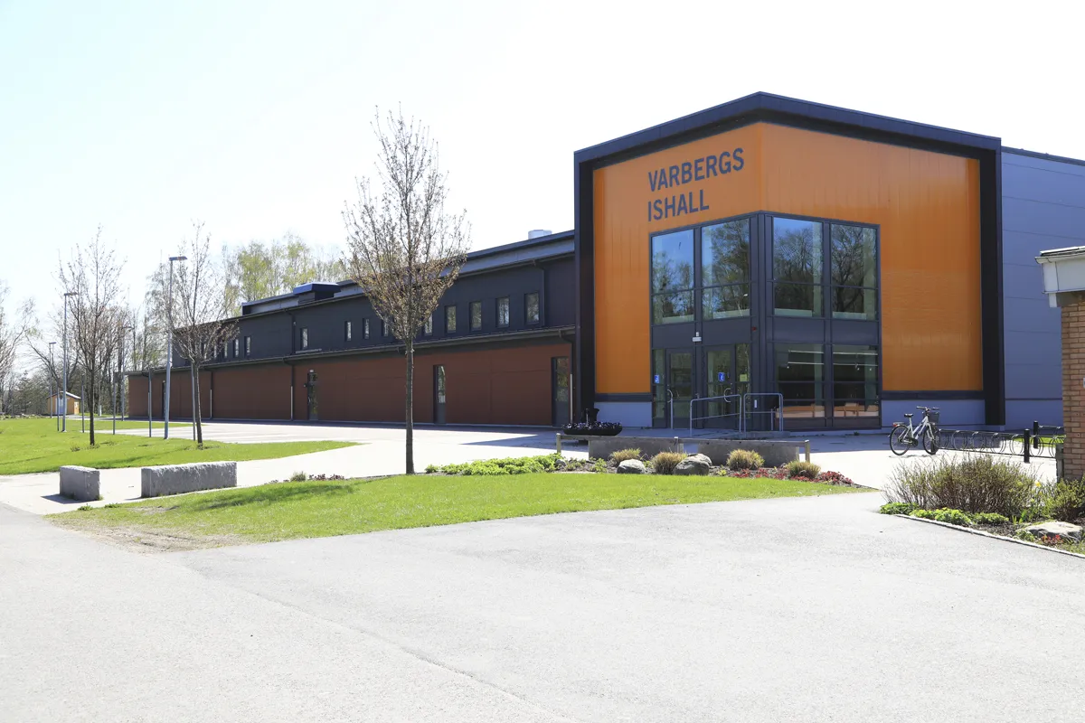 The front of Varbergs ishall, a dark steel building with an orange gable carrying the name, with a lawn and paved forecourt — Varbergs ishall (Pingvinen)