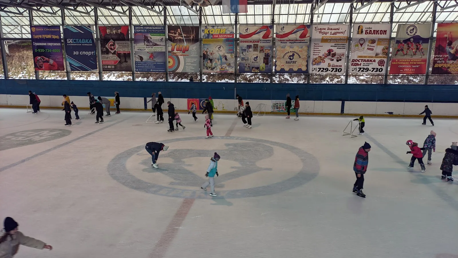 Skaters spread over the ice of the Varyag rink in Bryansk under the roof with its row of advertising banners along the far boards — Varyag Sports Park Ice Rink Bryansk