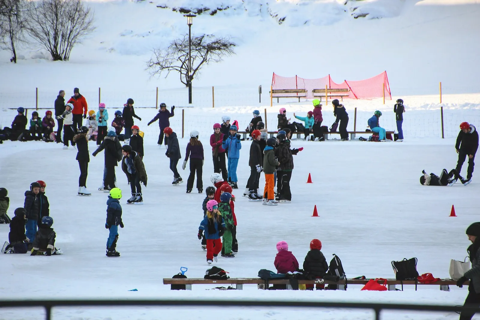 Barn och skridskoåkare på det snöpudrade isen på skridskoplanen i Vasaparken i Stockholm, med orange koner och en bänk vid kanten. – Vasaparkens skridskoplan