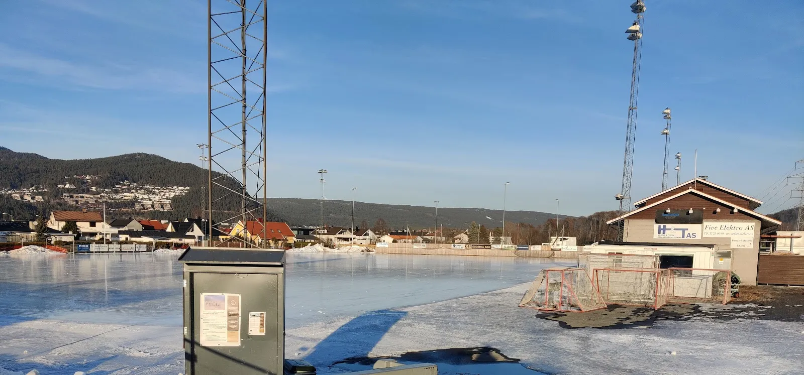 Vassenga-banen i dagslys, med klubbhuset, flomlysmastene og byen Mjøndalen på den andre siden av isen. — Vassenga kunstisbane