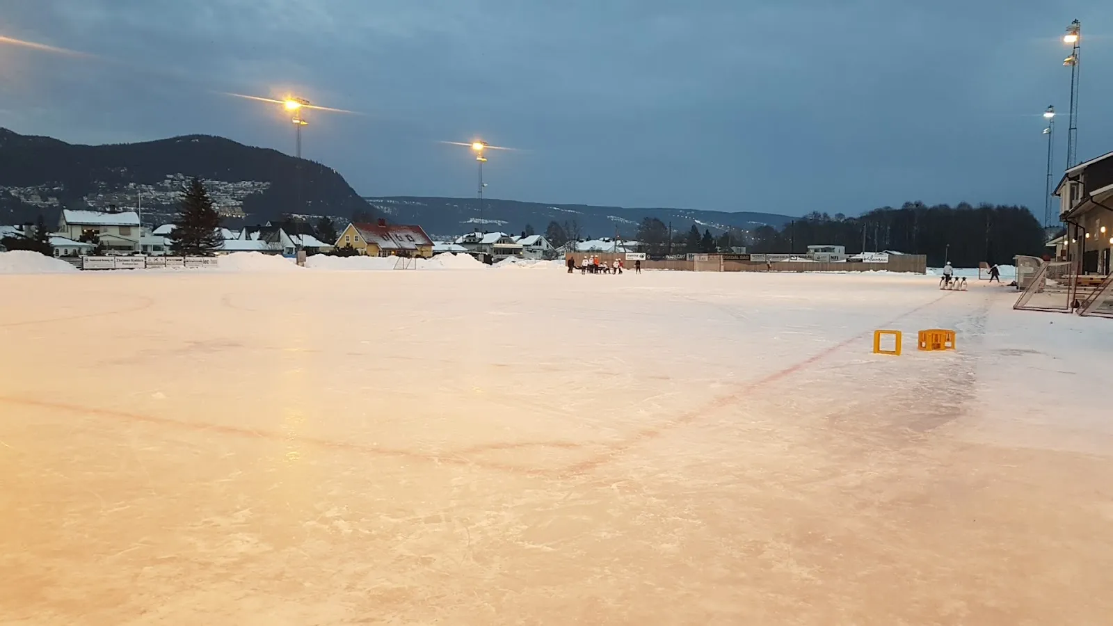 Utendørsisen på Vassenga i skumringen, med flomlys og åsene i Mjøndalen bak. — Vassenga kunstisbane