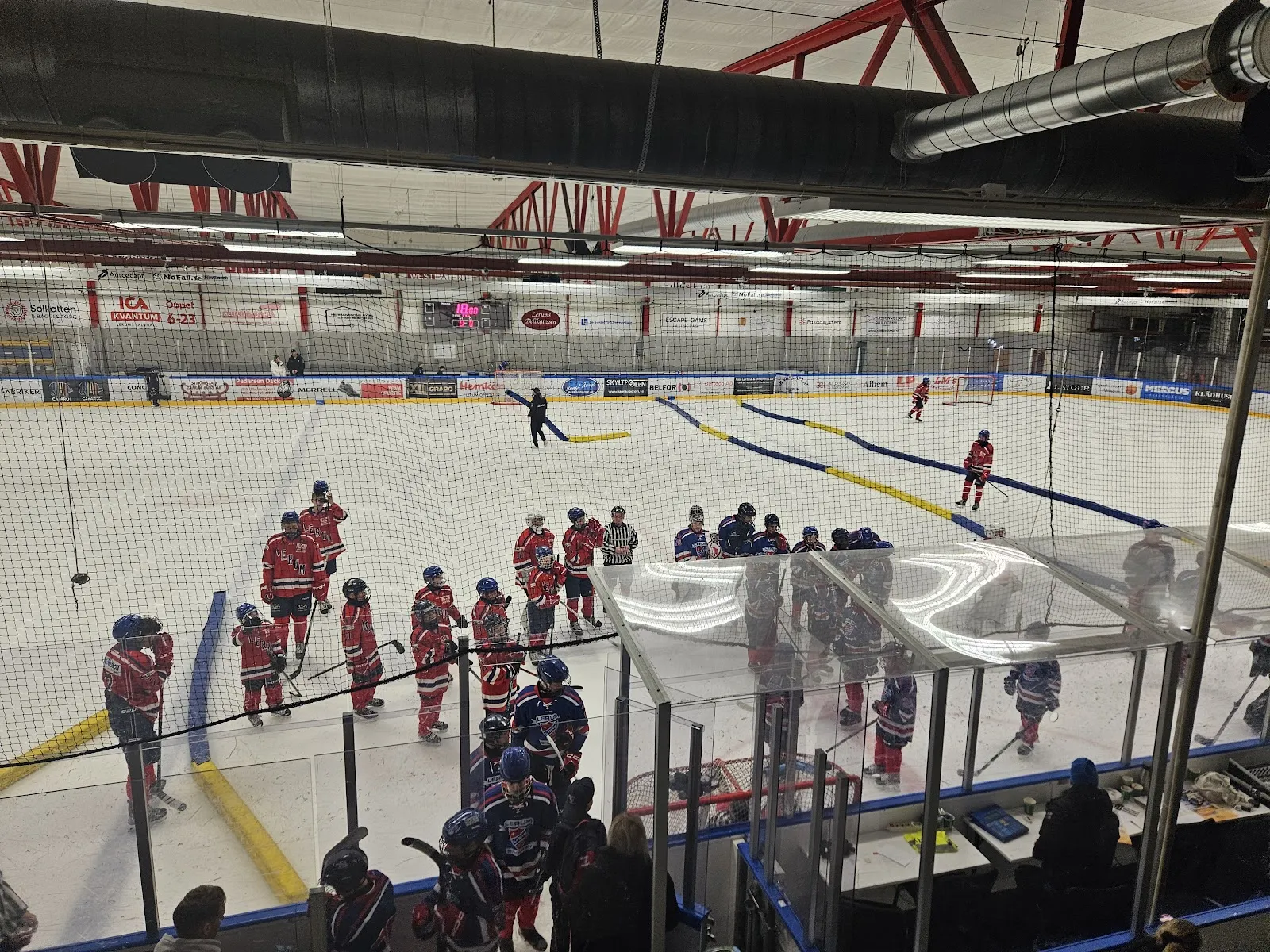 Vy ovanifrån över den inomhusbelägna isen med ungdomshockeyspelare och lagbänkarna längs sargen. – Vättlehallen ishall