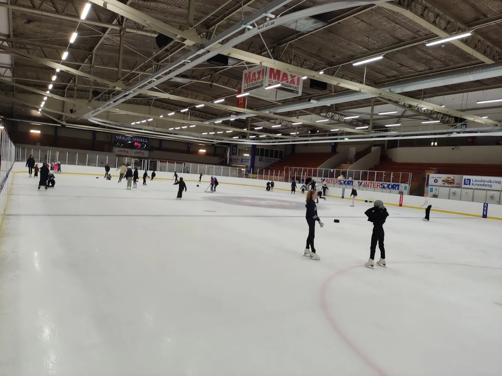 Skridskoåkare utspridda över isen i Växjö Ishall, sedda från sargen, med takbelysningen och den långa sidoläktaren bakom. – Växjö Ishall