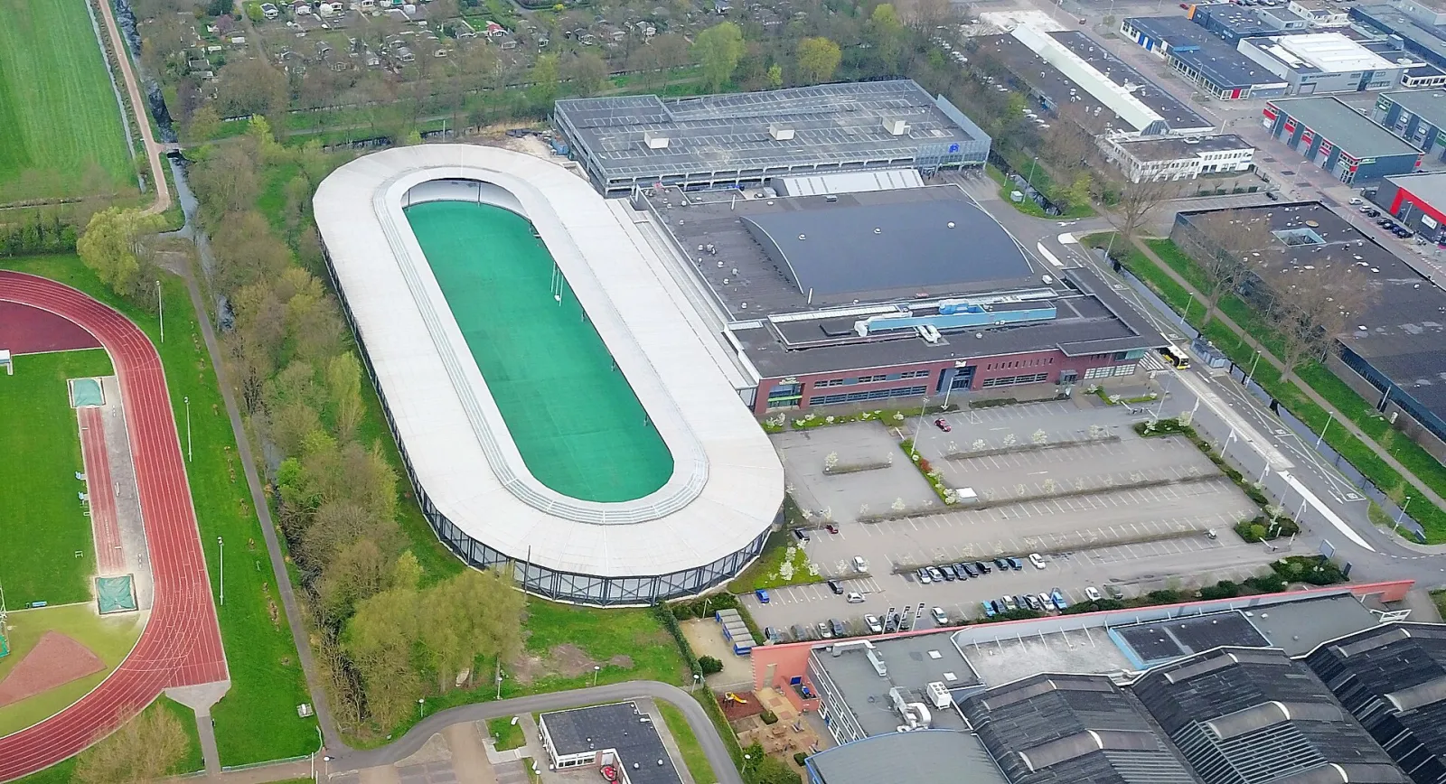 Luchtfoto van de Vechtsebanen: de half overdekte 400 m-ovaal met groen middenterrein, de ijshockeyhal en het entreegebouw naast de parkeerplaats – De Vechtsebanen