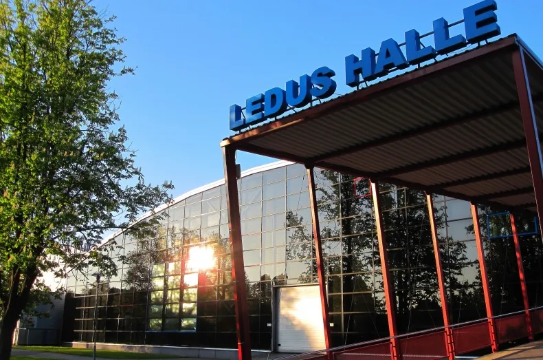 The Ventspils Ledus halle with its name above the glass front and a steel canopy, seen in low sun with trees beside it. — Ventspils Ledus halle