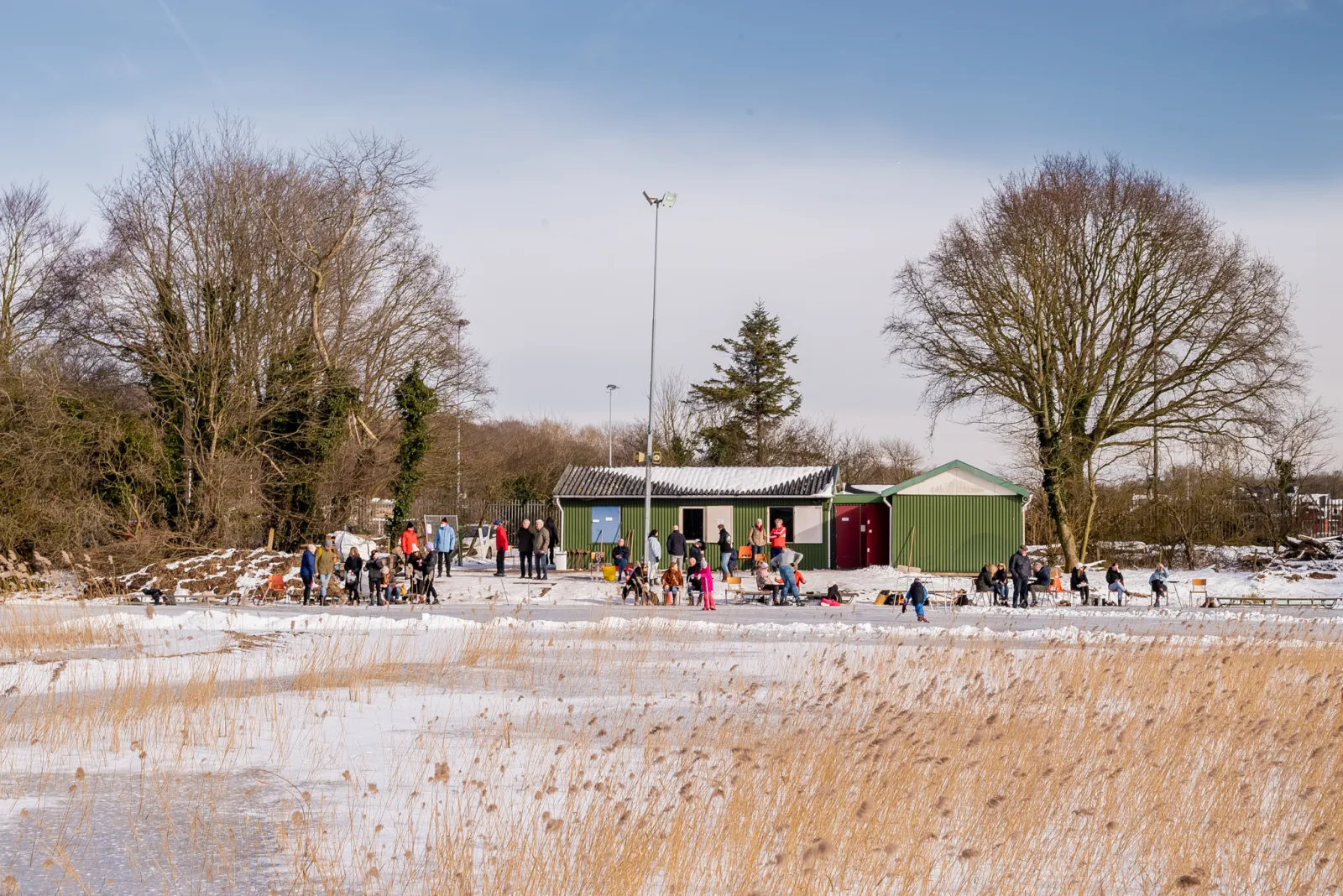 Skaters at a distance on the snow-dusted natural ice at Castricum aan Zee in February 2021, with the green clubhouse huts and bare trees behind the reeds. — Natuurijsbaan Castricum aan Zee (VKIJ)