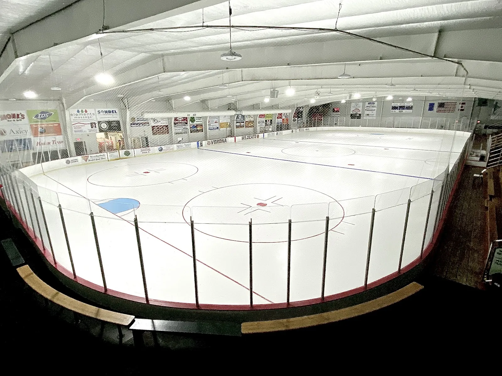 Toda la pista vacía de Verona Ice Arena, amplia vista de ojo de pez desde una esquina elevada con toda la pista, vallas de los VERONA WILDCATS, mantas de patrocinadores a lo largo de las paredes y un techo blanco aislado a dos aguas. — Verona Ice Arena