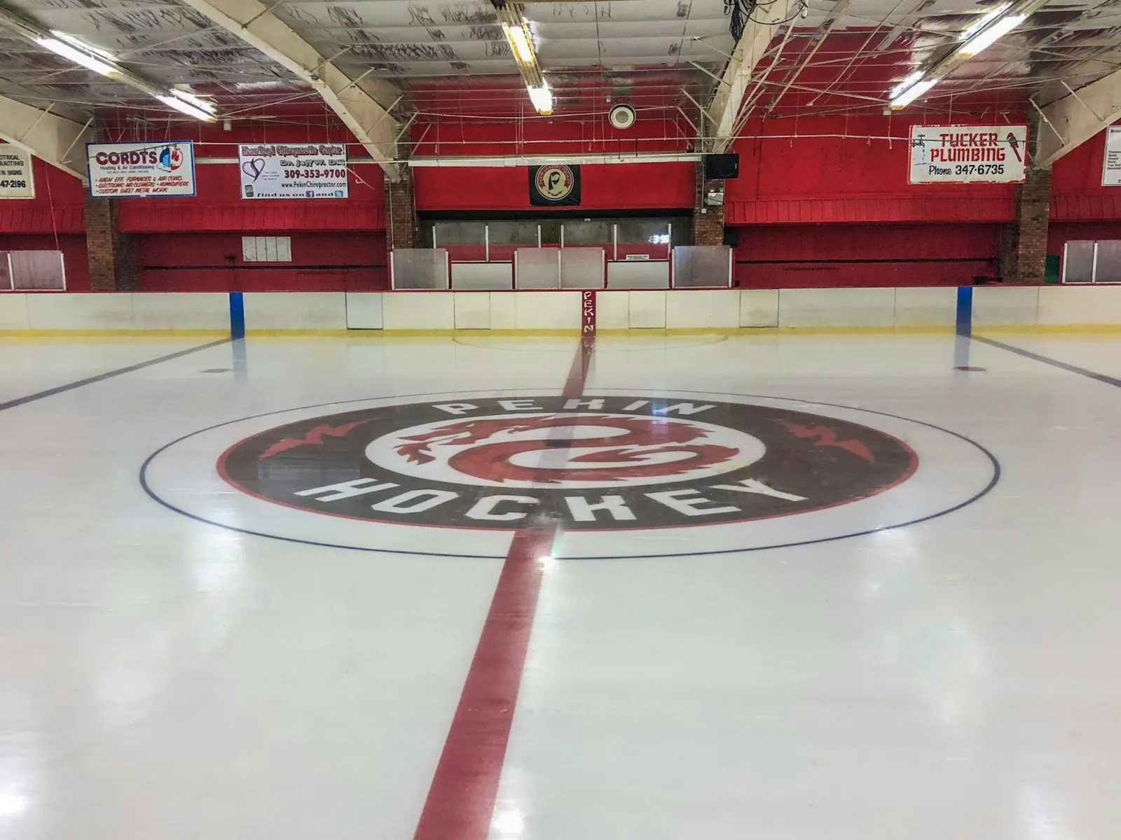 Empty ice sheet at center ice, showing the round 'Pekin Hockey' dragon logo, red dasher boards and bleacher fronts painted red, local sponsor banners (Cordts, Tucker Plumbing), and a Pekin hockey team banner hanging above the penalty boxes. — Veterans Memorial Arena (Pekin)