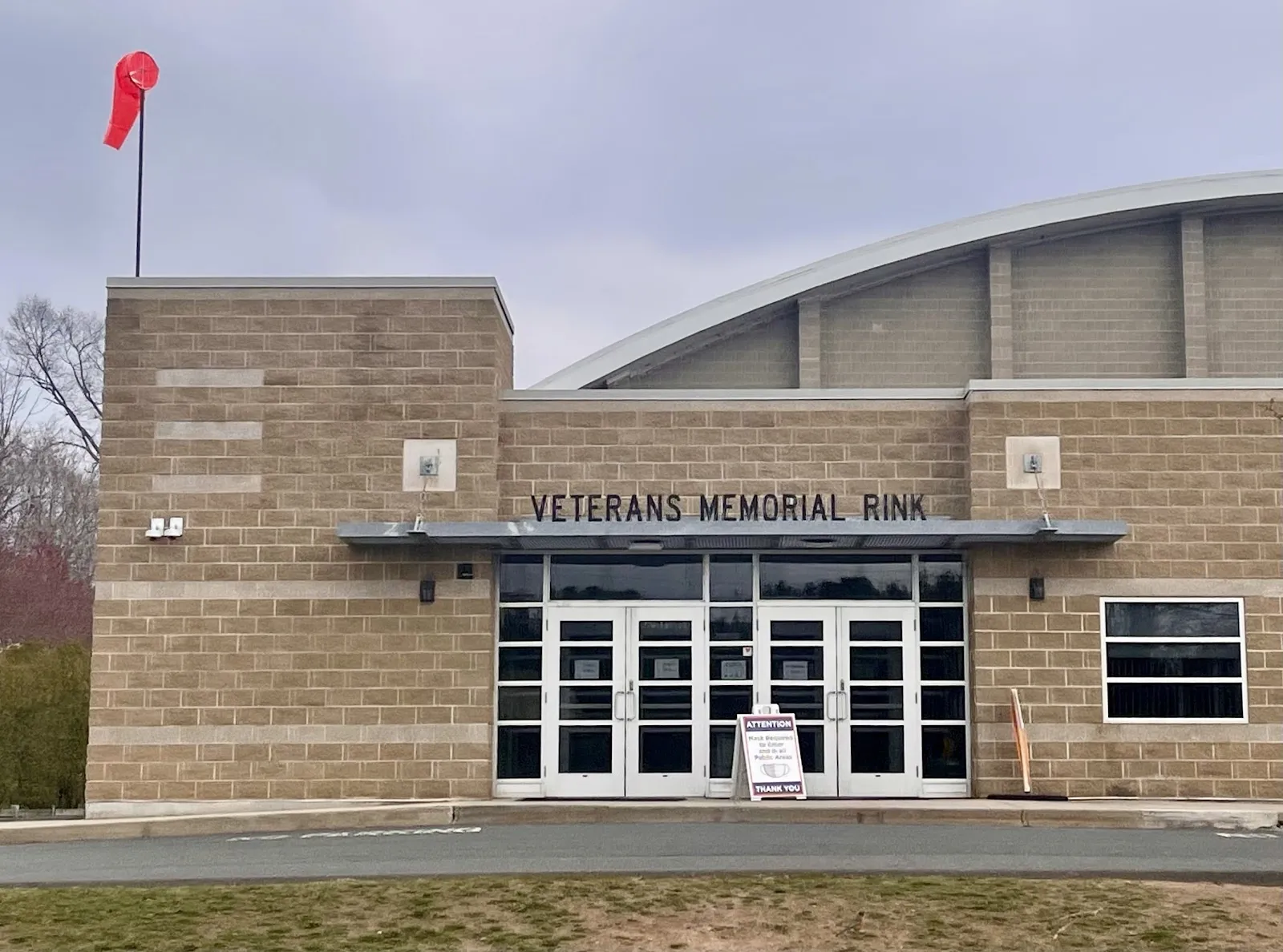 La entrada de ladrillo color arena de Veterans Memorial Rink, con su nombre sobre puertas de vidrio, un techo metálico arqueado que se eleva detrás y un atrio pavimentado al frente. — Veterans Memorial Skating Rink (West Hartford)