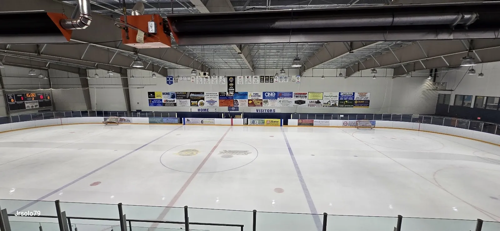 Una vista amplia a lo largo de la pista, con unos cuantos jugadores en el extremo opuesto bajo un techo del que cuelgan banderines de patrocinadores y una larga pared verde detrás del vidrio. — Veterans Memorial Sports Complex
