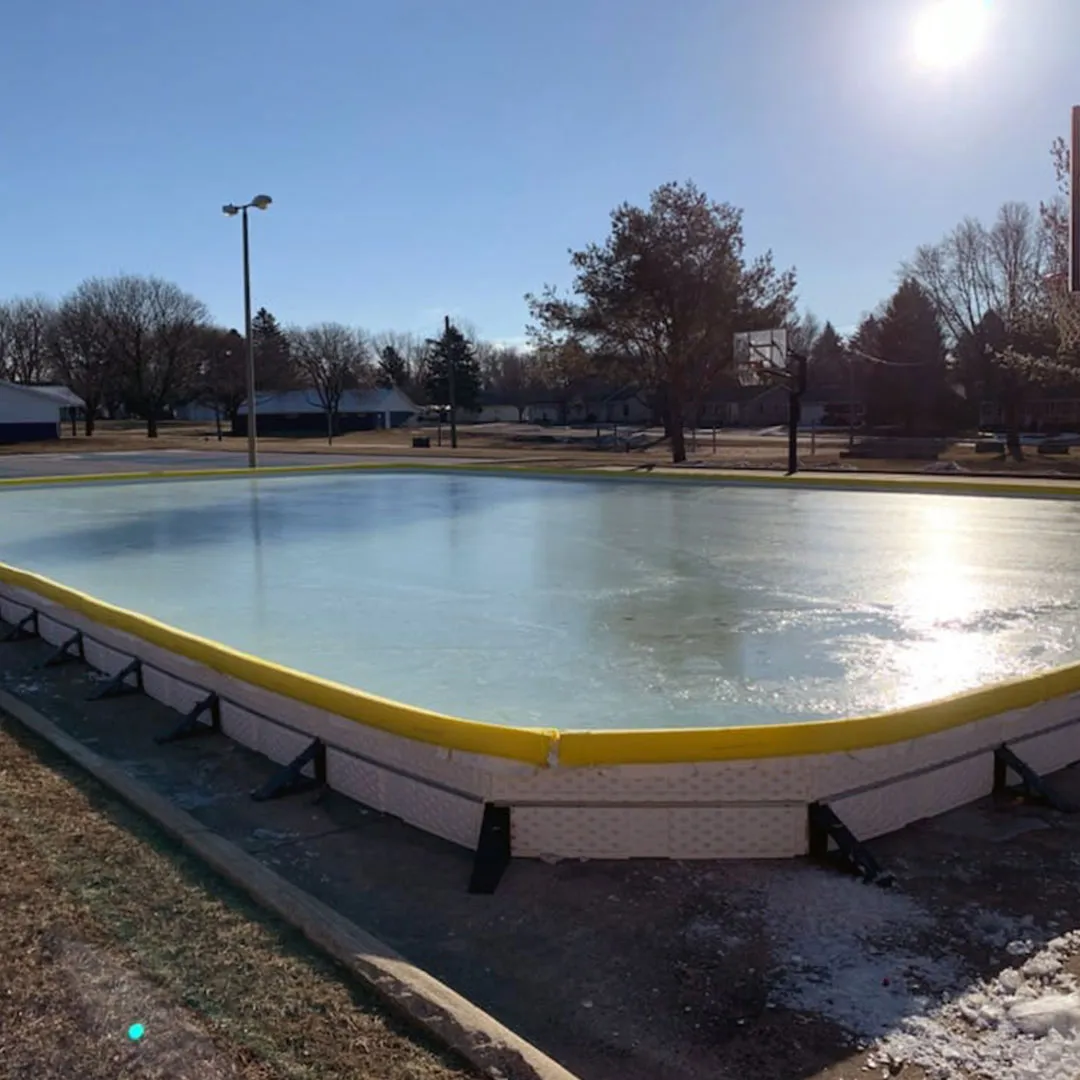 The boarded outdoor ice rink at Vets Park in Orange City on a sunny winter afternoon, a smooth sheet of ice inside yellow-topped white boards with bare trees and a light pole behind. — Vets Park Ice Rink