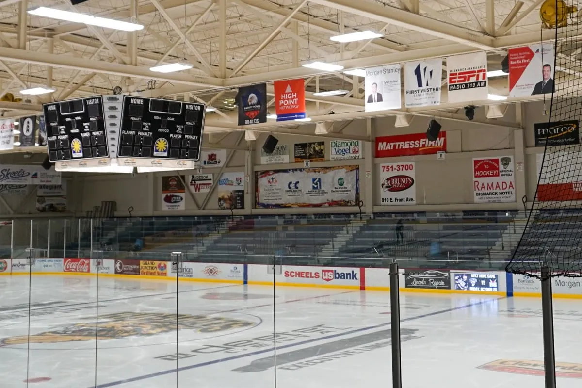 The empty Rink 1 ice sheet from behind the glass, with dasher-board and overhead sponsor banners (Scheels, US Bank, Ramada Bismarck Hotel, Bruno's Pizza) and empty bleachers. — VFW Sports Center